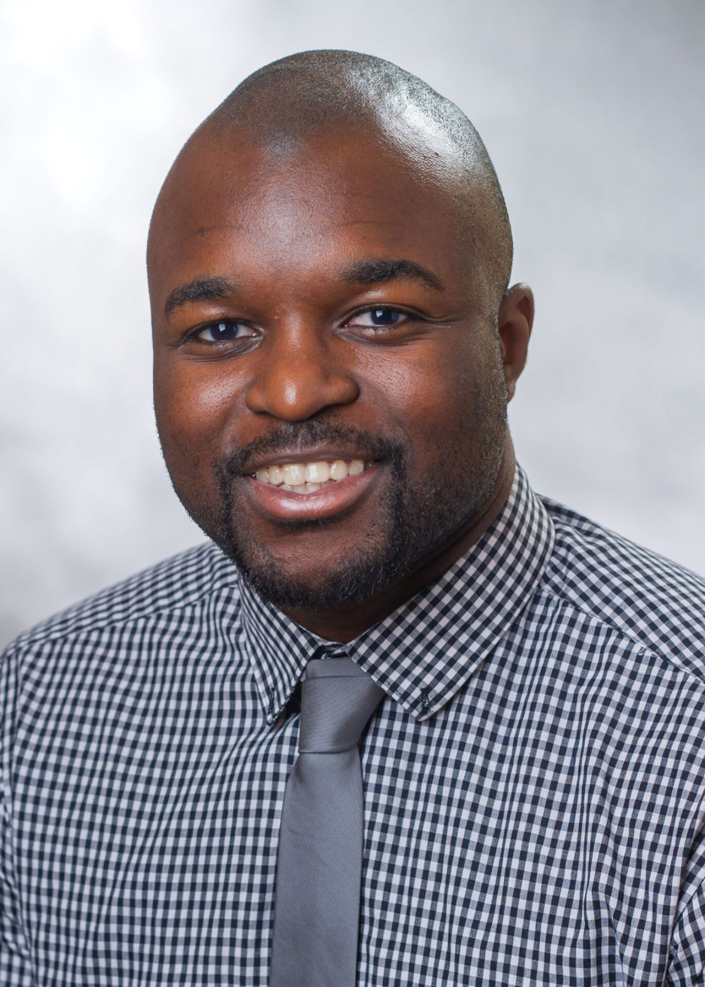 Professional headshot of a smiling man wearing a checkered shirt and a tie, with a clean-shaven head and a short beard, against a grey background.